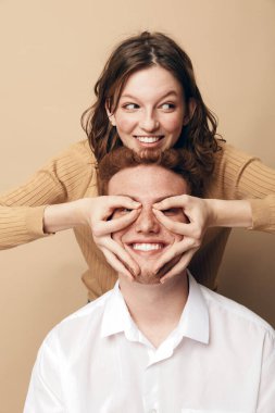 Gözleri kapalı gülümseyen adam, arkasından ellerle bardakları şekillendiren kadının neşeli hareketlerinden hoşlanır. Sıcak bej arka plan ve günlük tarz neşe, dostluk ve şefkat ifade eder.
