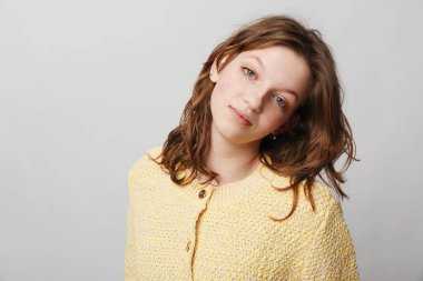 Gri arka planda sarı kazaklı, kahverengi saçlı genç bir kadının portresi. Basit bir stüdyo ortamında yumuşak ışıklandırma ve doğal makyajla huzurlu bir ruh hali sergiliyor..