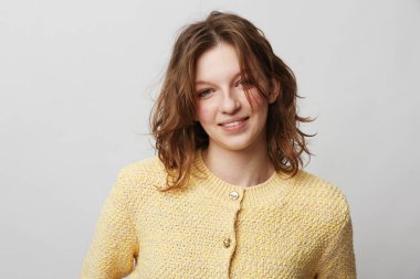 Sarı örgü süveter giymiş, düz bir arka plana hafifçe gülümseyen dalgalı kahverengi saçlı mutlu genç bir kadının portresi. Sıradan moda anlayışı, doğal güzellik ve pozitif ruh hali..