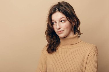 Bej balıkçı yaka kazaklı, kıvırcık saçlı, bej arka planda şüpheli ifadeli genç bir kadının portresi. Şüphe kavramı, merak, gündelik tarz ve duygu..