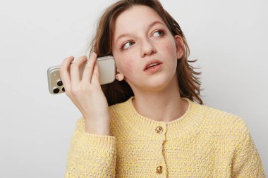 Sarı örgü kazak giyen genç bir kadının portresi akıllı telefonu kulağının dibinde tutuyor, düşünceli bir ifadeyle yana bakıyor. İletişim, teknoloji, gündelik yaşam tarzı ve yansıma kavramı.