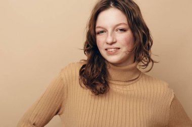 Tarafsız arka planda bej kazaklı, kıvırcık saçlı, gülümseyen genç bir kadının portresi. Doğal görünüş, gündelik moda ve yumuşak ışık stüdyoda sıcak ve rahat bir hava yaratır..