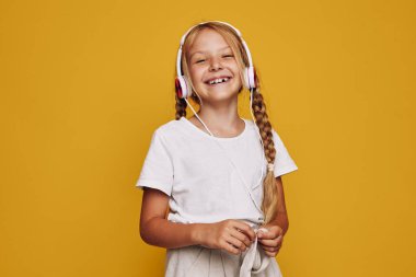 Uzun sarı örgülü gülümseyen genç kız parlak sarı bir arka planda beyaz kulaklıklı müzikten hoşlanıyor. Günlük beyaz tişört ve stüdyo çekimlerinde neşeli ifade..