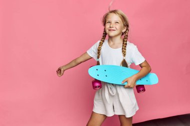 Saçları örgülü mutlu genç kız, pembe arka planda duran, sıradan beyaz bir elbiseyle mavi bir kaykay tutuyor. Stüdyo görüntüsü, çocukluk ve eğlence kavramı.