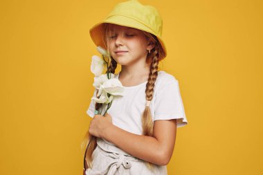 Sarı şapkalı ve beyaz tişörtlü gülümseyen genç kızın portresi göğsüne yakın parlak sarı arka planda beyaz çiçekler tutuyor. Çocukluk ve mutluluk kavramı.