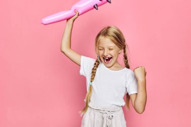 Beyaz tişörtlü heyecanlı bir kız kafasının üstünde pembe şişme bir oyuncak tutuyor. Gözleri kapalı ve neşeli bir ifadesi var. Parlak pembe arka planda stüdyo çekimi. Eğlence ve kutlama kavramı.