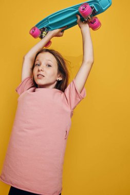 Pembe tişört giyen kendine güvenen genç bir kız canlı sarı bir arka planda turkuaz bir kaykayı kaldırıyor. Konsept gençliği, oyunculuğu ve sporu gösteriyor..