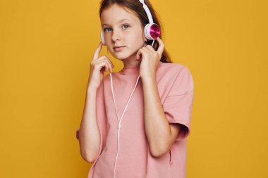 Pembe kazak giyen ve beyaz kulaklık takan genç kız katı turuncu bir arka plan önünde dikilirken dikkatle müzik dinliyor. Gençlik ve rahatlama kavramı.