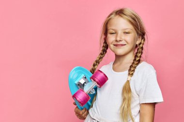 Mavi bir kaykayı tutan iki örgülü mutlu genç kız sağlam bir pembe arka planın önünde duruyor. Günlük beyaz tişört giyiyordu. Gençlik ve spor konsepti.