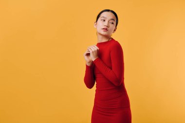 Dar kırmızı elbiseli genç bir kadın turuncu duvarın karşısında duruyor, düşünceli bakışlı eller, şık bir kıyafet ve temiz renkli bir stüdyo..