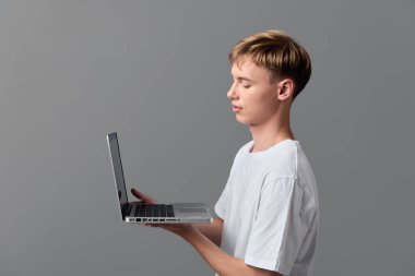 Beyaz tişörtlü bir çocuk dizüstü bilgisayar kullanıyor, ekrana odaklanıyor, basit bir arka planla dijital öğrenme ve teknoloji kullanımını gösteriyor.