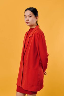 Kırmızı elbiseli şık bir kadın sarı arka planda poz veriyor. Siyah saçlı, kısa saçlı Asyalı bir manken. Sakin yüzüyle ve elleriyle kameraya bakıyor..