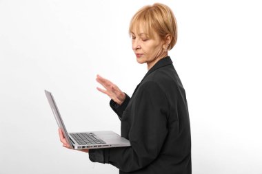 Ciddi orta yaşlı siyah ceketli bir kadının bir elinde dizüstü bilgisayar, diğerinde beyaz arka planda el hareketi yapması. İş ve teknoloji kavramı.