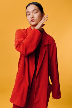Kırmızı takım elbiseli zarif bir kadın model canlı turuncu arka planda gözleri kapalı ve yüzünün yanında duruyor. Minimum makyajlı ve pürüzsüz saçlı çağdaş moda konsepti.