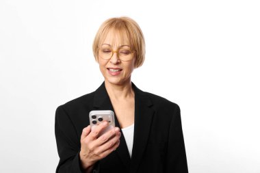 Gözlüklü, siyah ceketli, akıllı telefon ekranına bakarken gülümseyen orta yaşlı bir kadın. Beyaz arka planda izole edilmiş, dijital iletişim konsepti, teknoloji kullanımı ve günlük iş