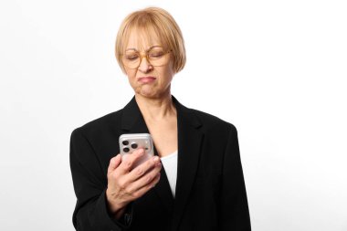 Gözlüklü, siyah ceketli mutsuz orta yaşlı kadın akıllı telefon ve somurtuyor. Beyaz arka plan stüdyo çekimi. Hayal kırıklığı, iletişim sorunları ve modern teknoloji kavramı.