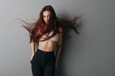 Gevşek saçlı, rahat beyaz bluz ve siyah pantolon giyen, moda ya da güzellik anlayışı için gri duvarın önünde gözleri kapalı poz veren kadın model..