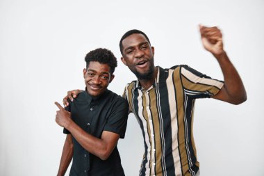 Siyah tenli Afro-Amerikalı erkekler birbirlerine yakın duruyorlar, gülümsüyorlar ve dostluk gösteriyorlar. Sıradan tarz, pozitif ruh hali ve beyaz, pürüzsüz arka planda rahat duruş..