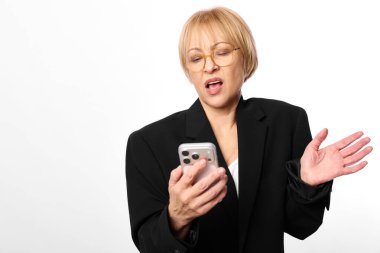 Kısa sarı saçlı, gözlüklü ve siyah ceketli şaşırmış bir kadın akıllı telefon ve beyaz arka planda el hareketleriyle hareket ediyor. Teknoloji, iletişim ve duygu kavramı.