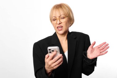 Kafası karışmış orta yaşlı, kısa sarı saçlı, gözlüklü ve siyah ceketli bir kadın akıllı telefonu elinde, diğer eli beyaz arka planda olan hareketler, hayal kırıklığı ve iletişim konsepti..