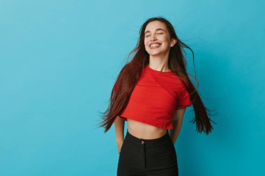 Kırmızı gömlekli ve siyah pantolonlu mutlu kadın gözleri kapalı ve gülümsüyor. Sade mavi arka planda, uzun saçlı, günlük kıyafetli neşeli bir kadın portresi..