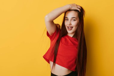 Saçları örgülü, kırmızı bluzlu, sarı arka planda duran, elleri kafasında gülümseyen ve kendine güvenen bir kadın model..