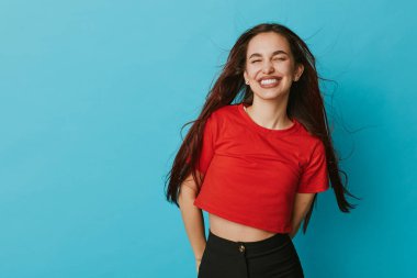 Kırmızı gömlekli ve siyah pantolonlu gülümseyen genç bir kadın mavi duvarın önünde poz veriyor. Uzun saçlı ve neşeli yüzlü bir kadın. Mutluluk ve kendine güveni kapalı mekanlarda ifade ediyor..