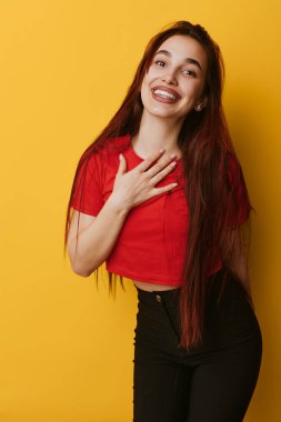 Uzun saçlı, kırmızı bluzlu ve siyah pantolonlu, sarı duvarın önünde duran gülümseyen genç bir kadın. Elini göğsüne koy, neşeli ruh hali, günlük tarz ve pozitif vücut dili..
