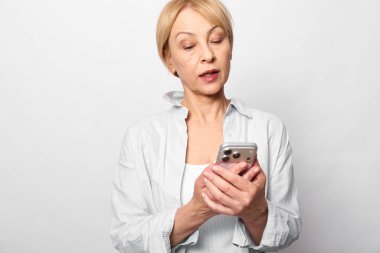 Beyaz gömlek giyen olgun bir kadının elinde iki eliyle beyaz arka plandaki ekrana bakan bir akıllı telefon var. Mobil teknoloji, iletişim ve duygusal tepki kavramı.