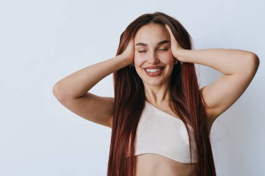 Gözleri kapalı gülümseyen ve elleri beyaz tişörtlü genç bir kadın. Uzun saçlı, mutlu bir kadın stüdyoda poz veriyor. Hafif ışıklandırma ve minimalist bir altyapısı var..