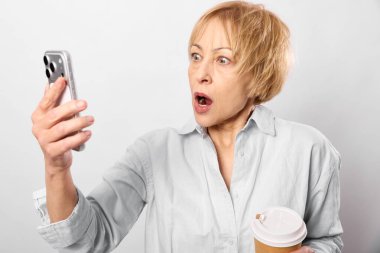 Beyaz gömlek giyen, kısa saçlı, şok olmuş olgun bir kadın elinde akıllı telefon ve kahve fincanı ile ekranda şaşkın şaşkın bakıyor. Beyaz arka planda izole edilmiş. Tepki ve iletişim kavramı.
