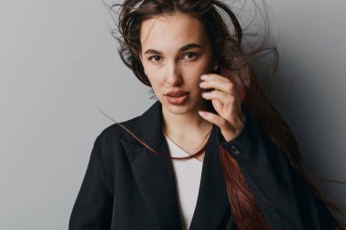 Siyah ceketli, beyaz gömlekli, uzun dalgalı saçlı genç bir kadının portresi. Stüdyoda, yumuşak ışıklandırma ve doğal ifade ile, düz gri arkaplanda el üstünde duran kadın model..