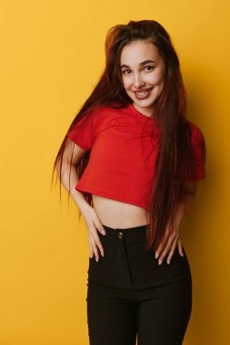Uzun saçlı, kendine güveni tam bir genç kadın kırmızı bluz ve siyah pantolon giyiyor. Stüdyoda sarı arka planda gülümseyerek poz veriyor. Sıradan moda portresi için..
