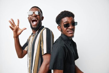 Siyah tenli, günlük giysiler ve güneş gözlüklü Afro-Amerikalı erkekler beyaz stüdyo arka planında özgüvenli ve mutlu ruh halleriyle sırt sırta dururlar..