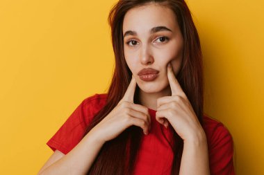 Düz saçlı, kırmızı gömlekli, yanaklarında parmakları olan genç bir kadının portresi. Doğal makyajlı ve parlak sarı arkaplan önünde ciddi yüzlü bir kadın model..