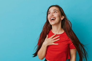 Uzun kahverengi saçlı, kırmızı bluzlu, gülümseyen ve göğsüne dokunan mutlu bir kadın mavi arka planda yukarı bakıyor. Neşeli bir kadının portresi neşe ve minnettarlık gösteriyor..