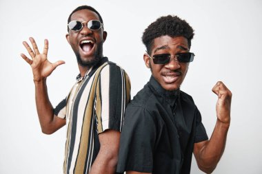 Siyah tenli, günlük kıyafetler ve güneş gözlüğü takmış, sırtüstü gülümseyen, neşeli ve enerjik pozlar veren Afro-Amerikalı erkekler stüdyoda beyaz arka planda..