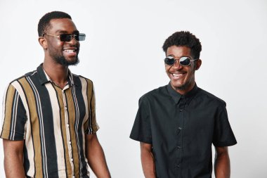 Siyah tenli, güneş gözlüklü ve beyaz arka planda poz veren iki siyah adam. Arkadaşlık, mutluluk ve stil. Olumlu ruh hali ve modern moda..