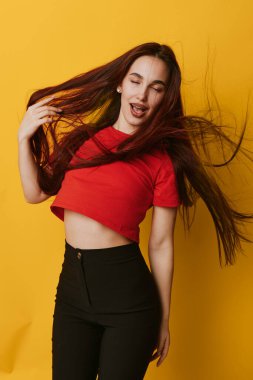 Kırmızı gömlekli, siyah pantolonlu, uzun saçlı, gözleri kapalı, sarı arka planda duran bir manken. Gençlik, yaşam tarzı, hareket, güven ve modern günlük kıyafetler..