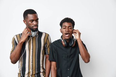 Koyu tenli genç Afro-Amerikalı erkekler müzikten hoşlanıyor, biri kulaklıkları ayarlıyor ve diğeri şarkı söylüyor, beyaz arka planda çizgili ve siyah gömlekli gündelik kıyafetler..