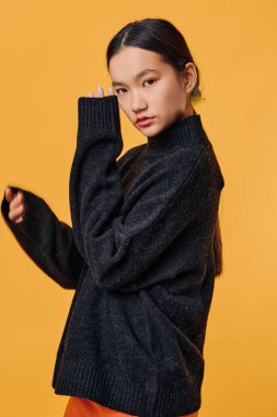 Parlak turuncu arka planda duran siyah kazaklı Asyalı kadın. Moda portresi, doğal makyajlı ve uzun siyah saçlı, elleri yüze yakın ve yumuşak ifadeli..