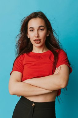Kırmızı bluzlu, siyah pantolonlu, çapraz kollu, kendine güvenen genç bir kadın mavi arka planda doğal ifade ve günlük tarzla duruyor..