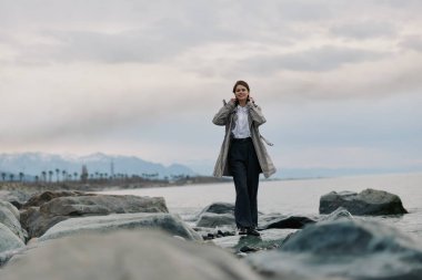 Ceket giyen ve günlük kıyafet giyen kadın bulutlu gökyüzünün altında sakin deniz kıyısında duruyor. Seyahat kavramı, doğa macerası, açık hava aktivitesi ve uzak dağları olan sakin sahil atmosferi..