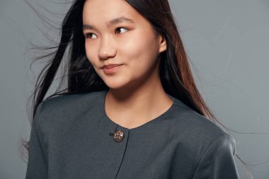 Siyah uzun saçlı, gri elbiseli, başı yana dönük poz veren genç bir kadın. Sade gri arka planda duran sakin yüzlü Asyalı kadın..