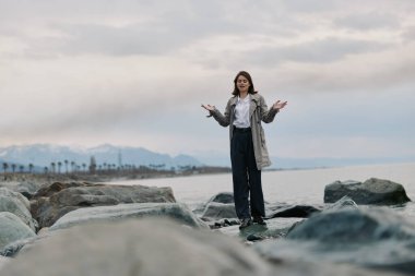 Sıradan giyinmiş bir kadın, bulutlu gökyüzü ve dağ sıraları olan sakin okyanusun yanındaki kayalarda poz verir. Doğa, moda ve açık hava yaşam tarzı kavramı.