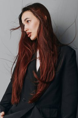 Siyah ceketli, dalgalı kızıl saçlı genç kadın gri desenli duvarın yanında duruyor. Modern moda konseptinde sakin yüzlü ve az makyajlı şık bir kadın modeli..