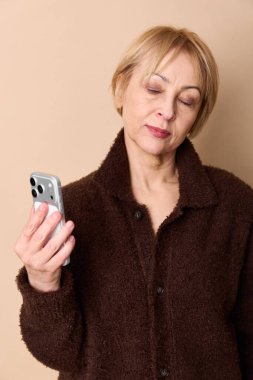 Koyu kahverengi ceketli, yorgun orta yaşlı bir kadın akıllı telefon tutar, gözleri yorgunluktan kapalıdır. Yorgunluk, stres, teknoloji yorgunluğu ve günlük dijital zorluklar kavramı.