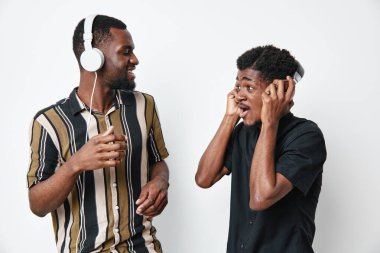 Koyu tenli iki siyah adam kulaklıkla müzik dinliyor, gülümsüyor ve günlük kıyafetlerle etkileşime giriyorlar. Genç arkadaşlar beyaz stüdyo arka planında ses ve eğlencenin keyfini çıkarıyorlar.