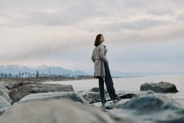 Paltolu bir kadın okyanusun kıyısındaki kayalarda durup bulutlu bir gökyüzünün altındaki ufka dikkatlice bakıyor. Doğal ortam barışçıl ve yansıtıcı bir hava yaratır..