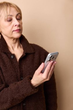 Kahverengi kazaklı, odaklanmış olgun bir kadın akıllı telefon tutuyor ve bej arka plandaki ekrana bakıyor. Orta yaşlı teknoloji kullanımı, dijital iletişim ve günlük yaşam kavramı.